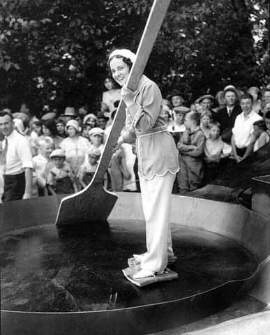 Woman with slabs of bacon tied to her feet standing in a giant skillet holding an enormous wooden spatula and smiles at the crowd, Chehalis, Washington