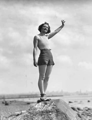 Woman in a swimsuit waving while standing on a large rock on a beach, probably Washington State